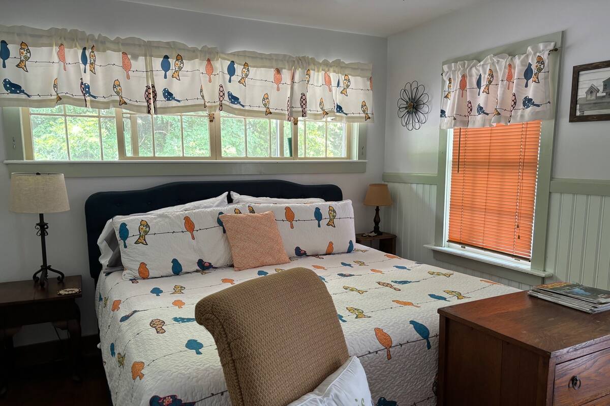 A bedroom with a bed, two bedside tables, a chair, a dresser, and a window with blinds.