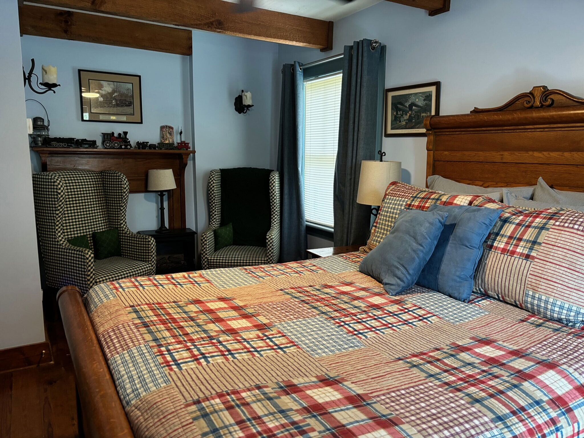 A bedroom with a plaid bedspread, two checkered armchairs, a wooden headboard, a nightstand with a lamp, and a window with curtains.