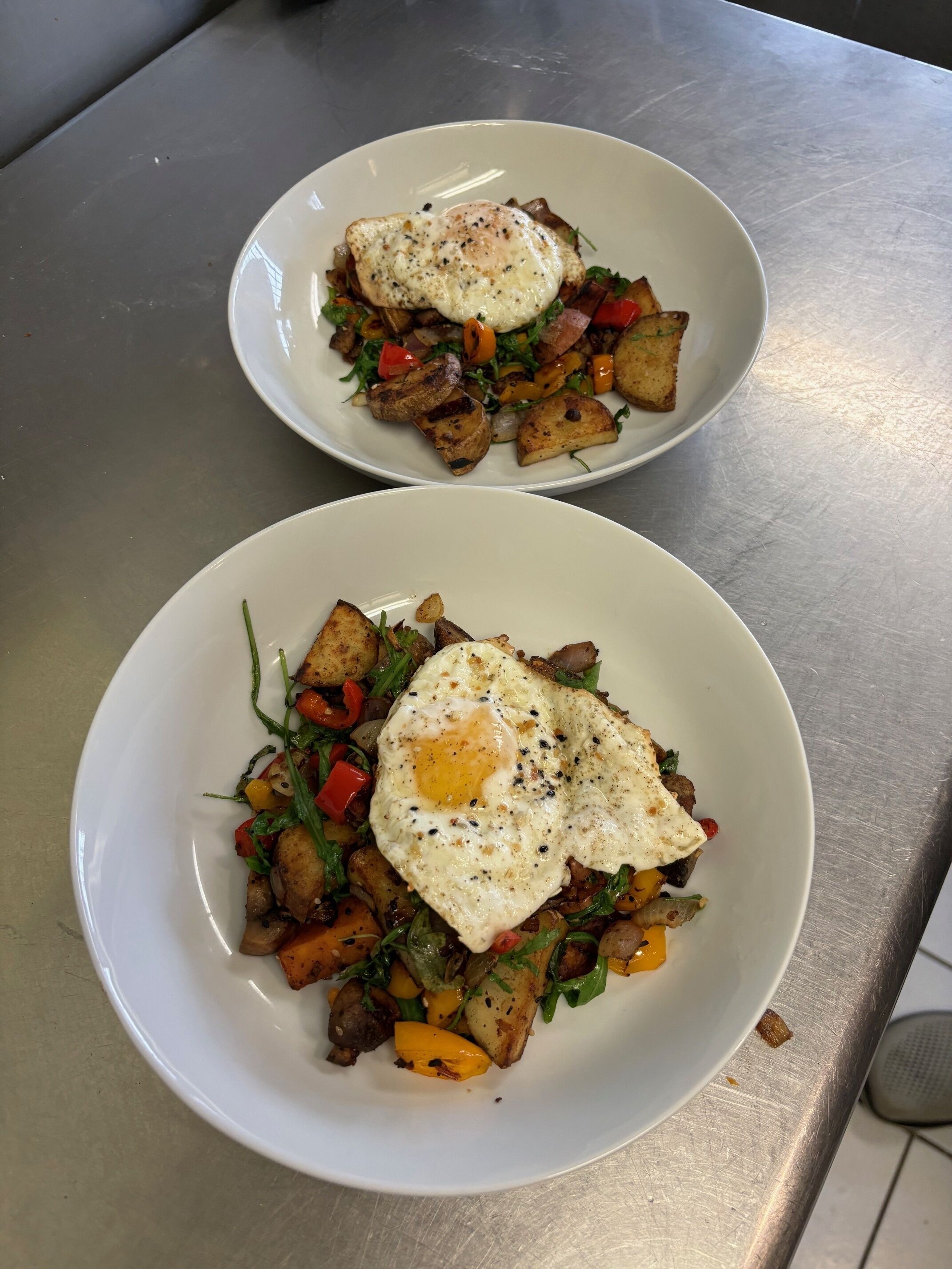 Two white plates with fried eggs and roasted vegetables on a stainless steel table.