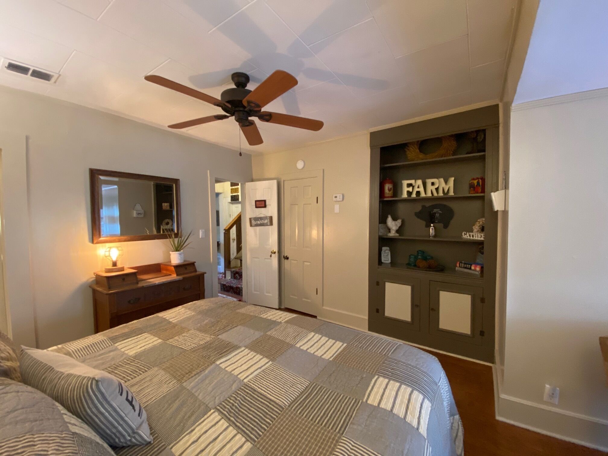 Une chambre à coucher comprenant un lit, un ventilateur de plafond, une commode, un miroir, une étagère avec des objets décoratifs et une porte donnant sur une autre pièce.