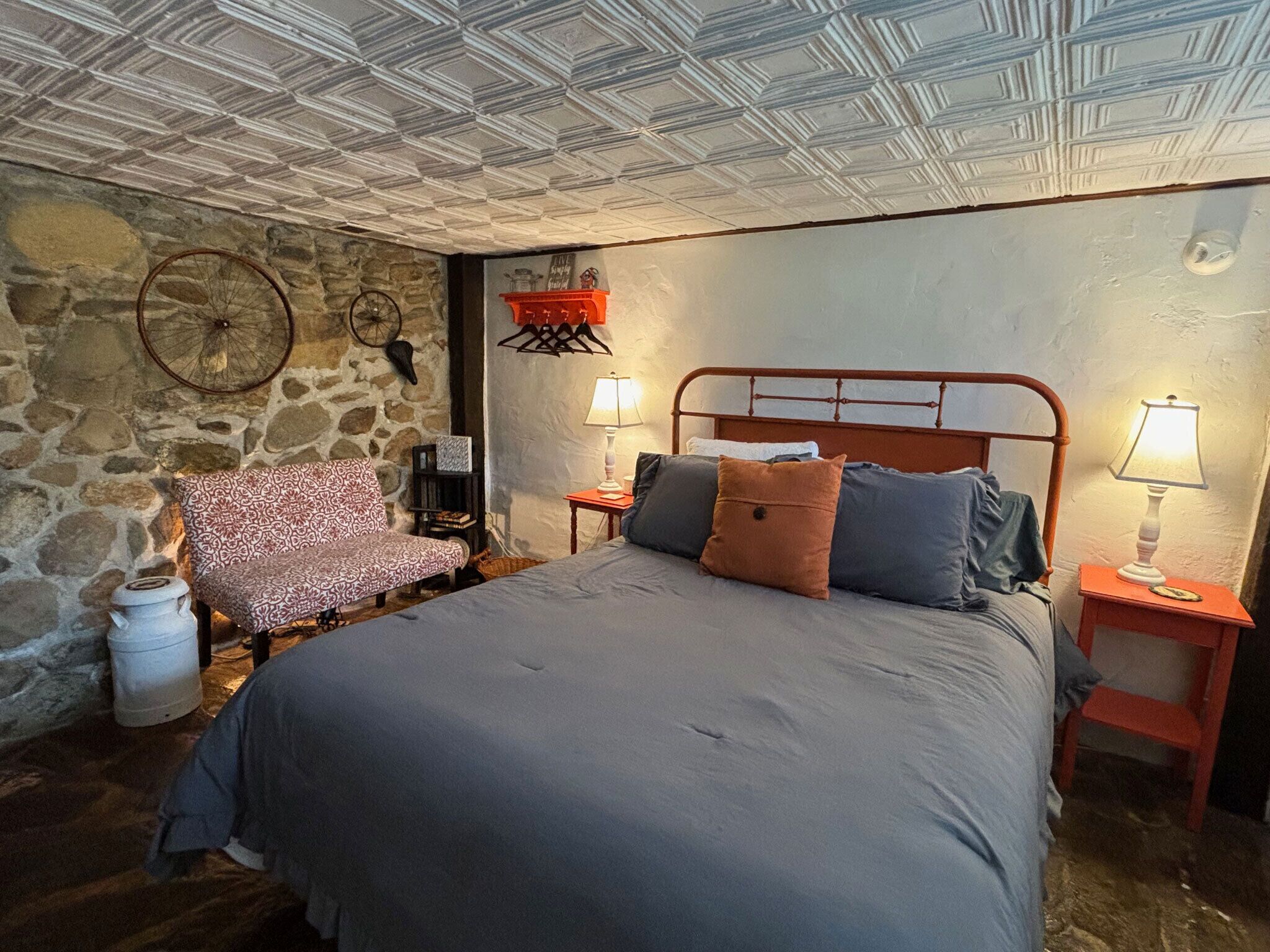 Un dormitorio con pared de piedra, un sillón estampado, una cama con muchos almohadones y dos mesitas de noche con lámparas.