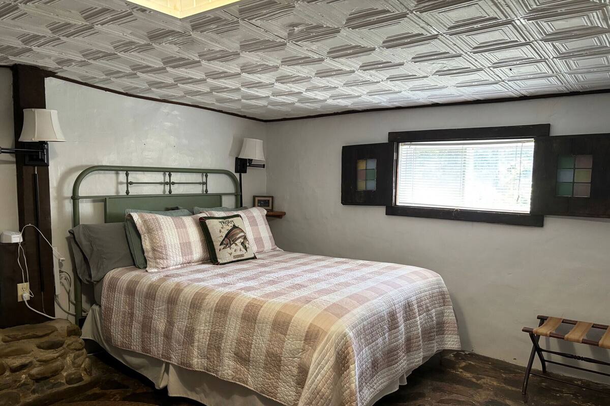 A bedroom with a bed, a nightstand, a lamp, a window, and a stone wall.