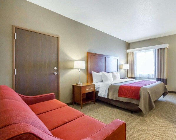 A hotel room with a bed, a nightstand, a lamp, a red sofa, and a door.
