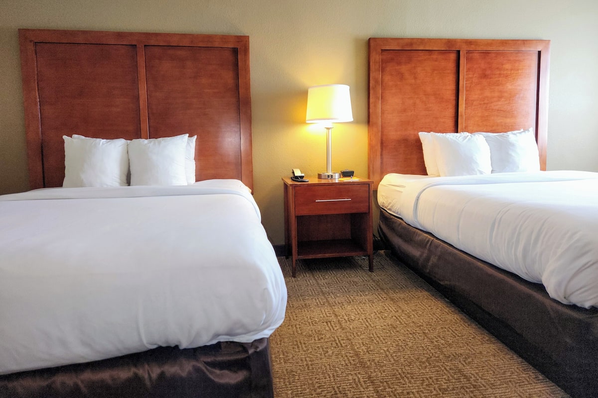 A hotel room with two beds, each with white bedding and wooden headboards. There is a nightstand with a lamp and a phone between the beds.