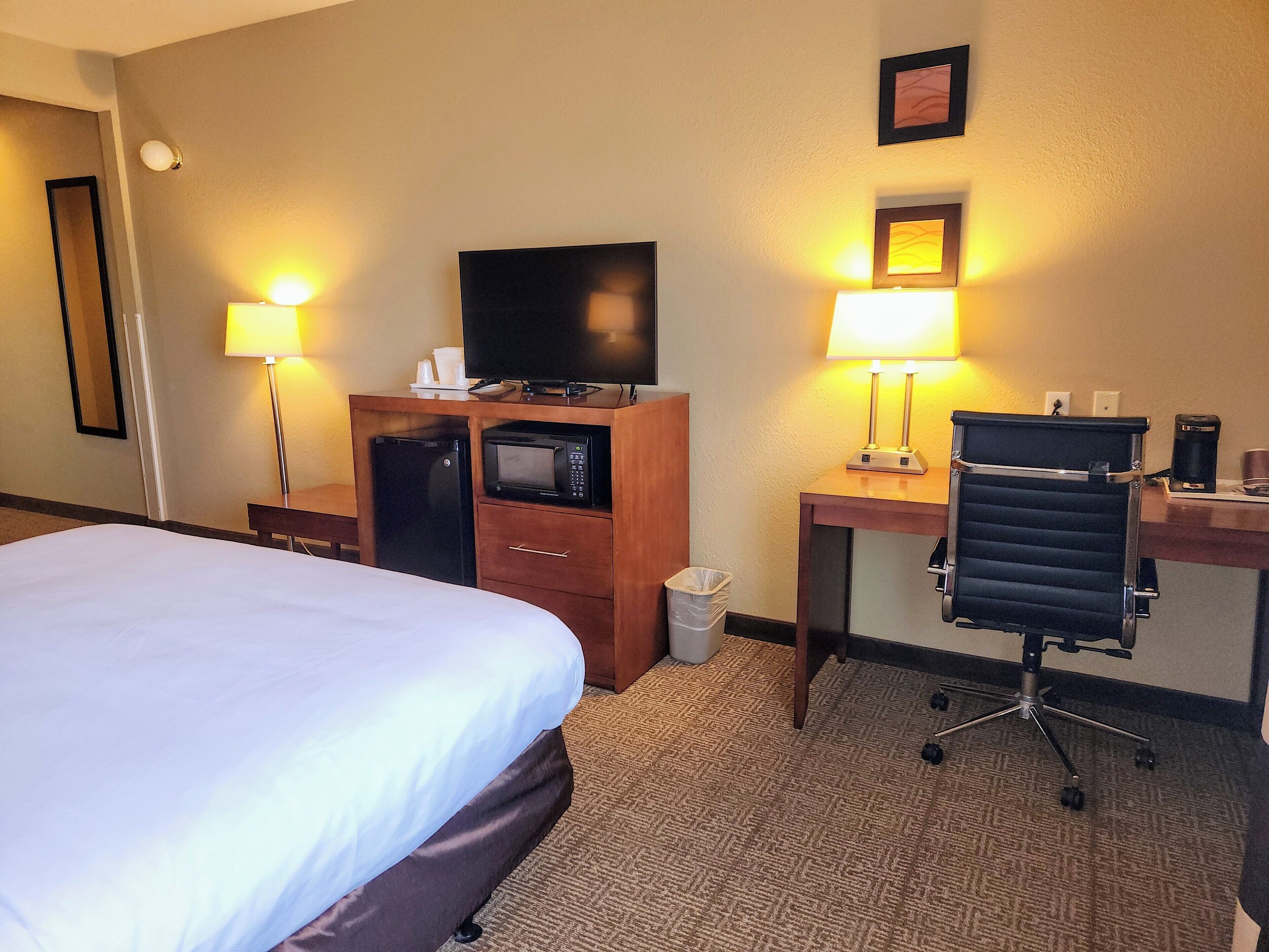 A hotel room with a bed, a desk with a computer, a TV, a microwave, a trash bin, and a chair.