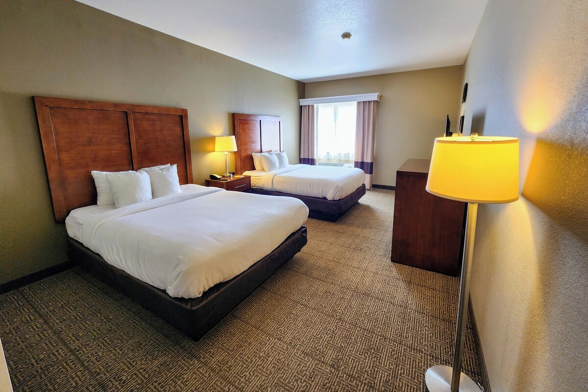 A hotel room with two beds, a nightstand, a lamp, and a window with curtains.