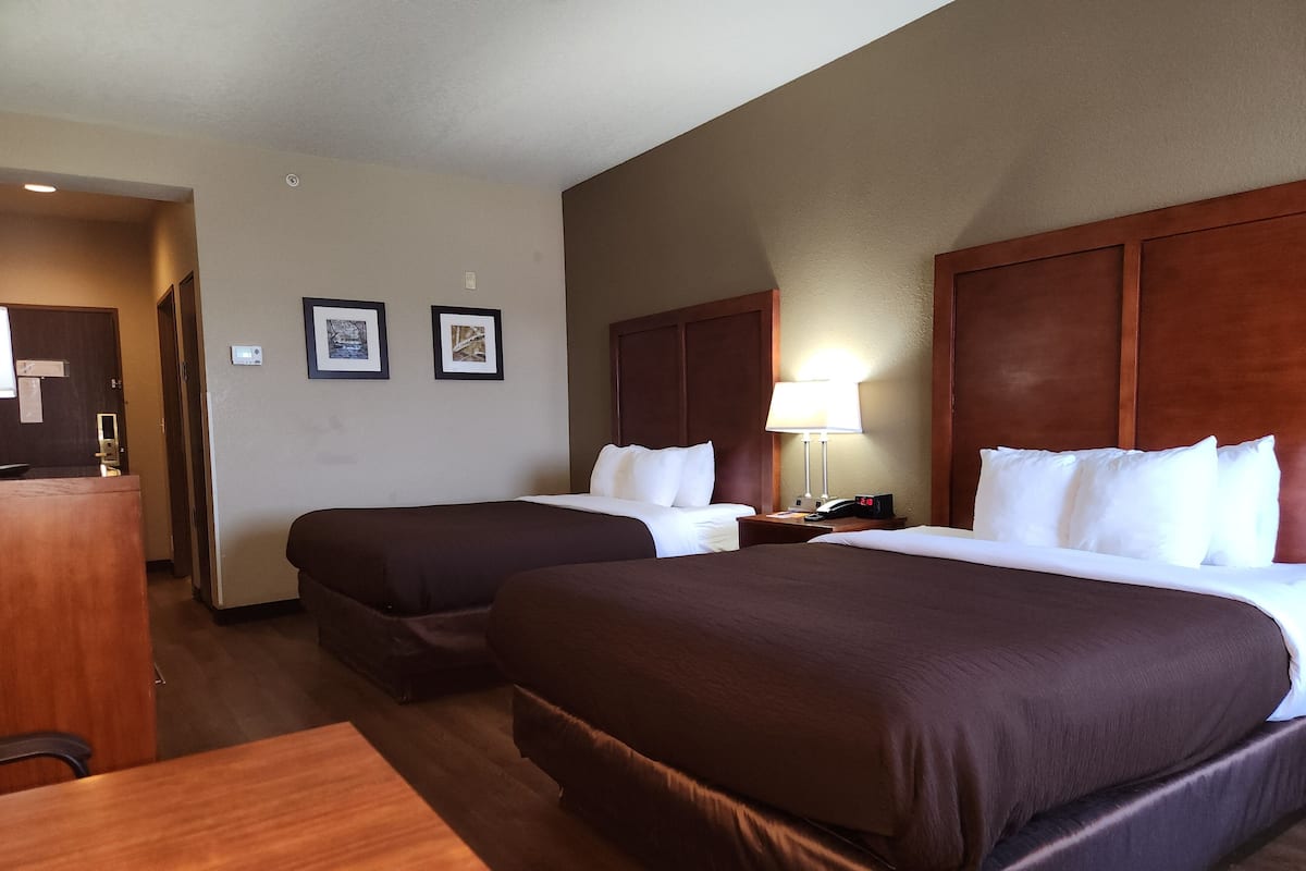 A hotel room with two beds, a desk, a lamp, and framed pictures on the wall.