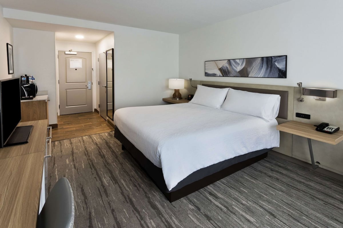 A hotel room with a bed, bedside table, lamp, and a painting on the wall.