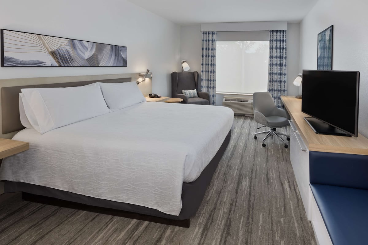 A hotel room with a large bed, a desk with a TV, a chair, a desk lamp, and a window with curtains.