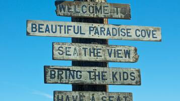 A wooden signpost with multiple directional signs, including "Welcome to Paradise Cove," "Sea the View," "Bring the Kids," "Have a Seat," and "Enjoy Malibu." A seagull is perched on top of the signpost.