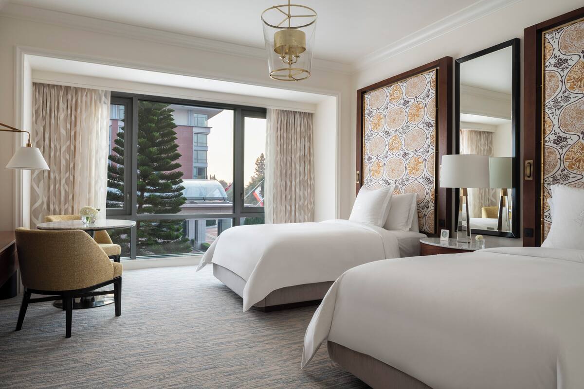 A hotel room with two beds, a sitting area with a chair, a table, and a large window with a view of a building and a tree.