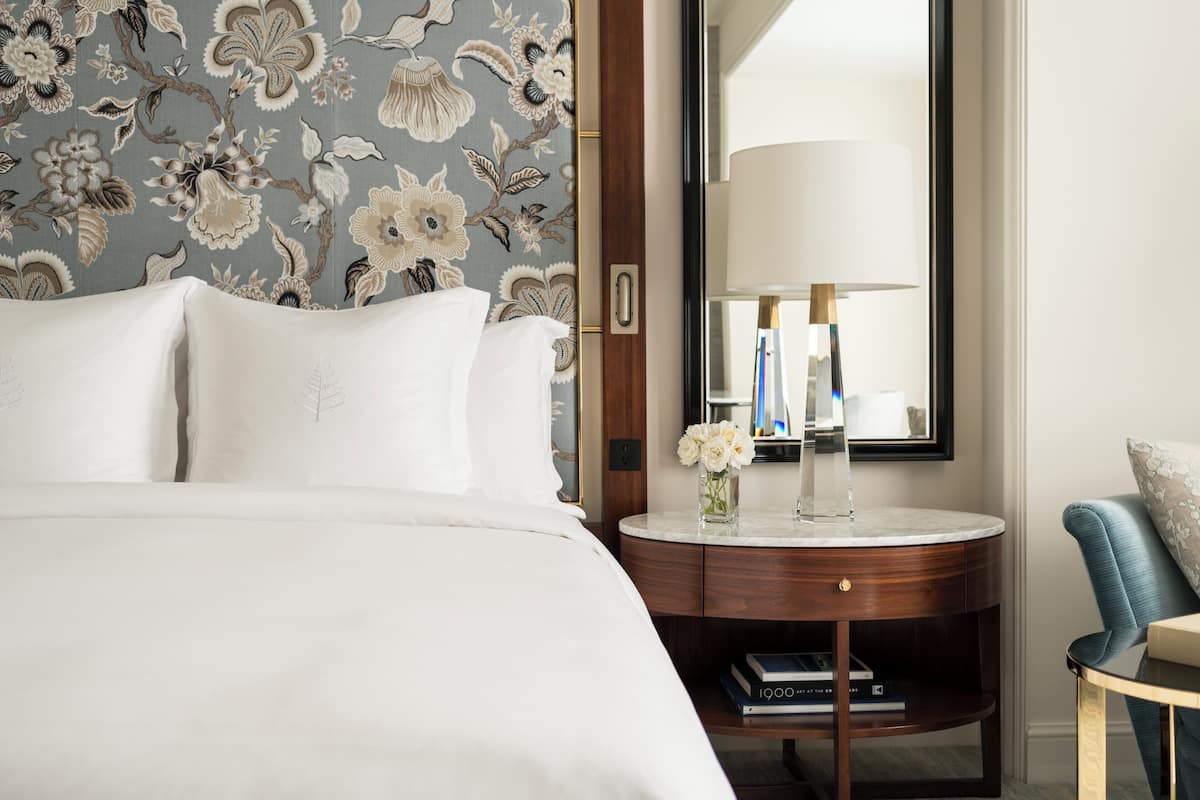 A neatly made bed with white linens, a round wooden nightstand with a vase of flowers, and a blue upholstered chair.