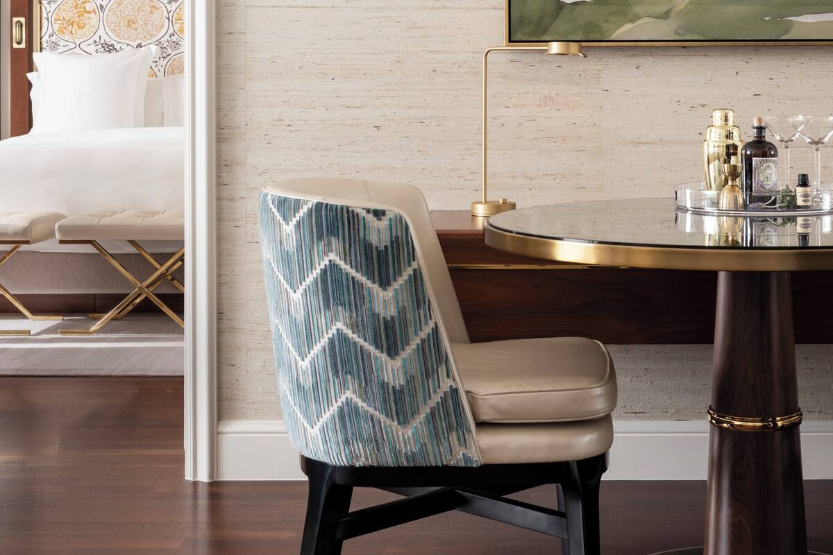 A modern hotel room with a large abstract painting, a patterned wallpaper, and a dining area with a round table and a chair with a chevron pattern.