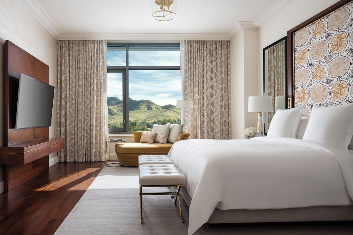 A modern hotel room with a large bed, a TV, a sofa, and a view of the hills.