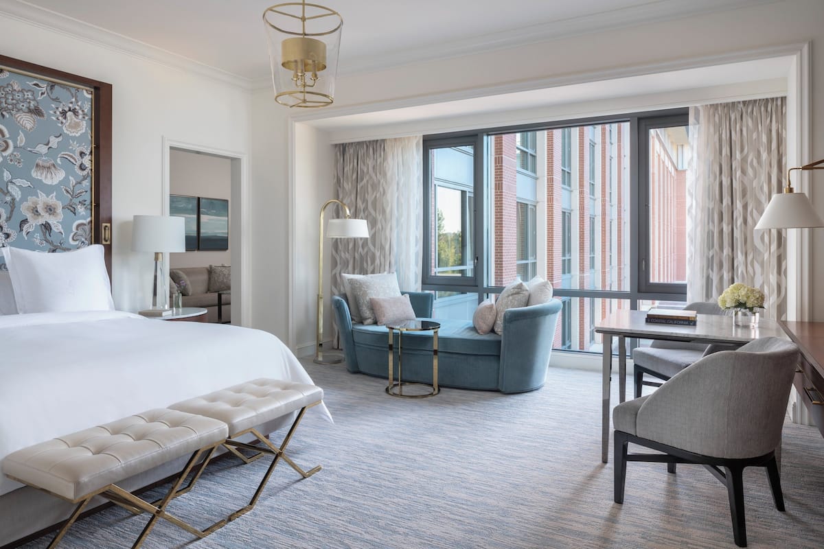 A hotel room with a large bed, a seating area with a sofa and armchairs, a dining table, and a view of the city through large windows.