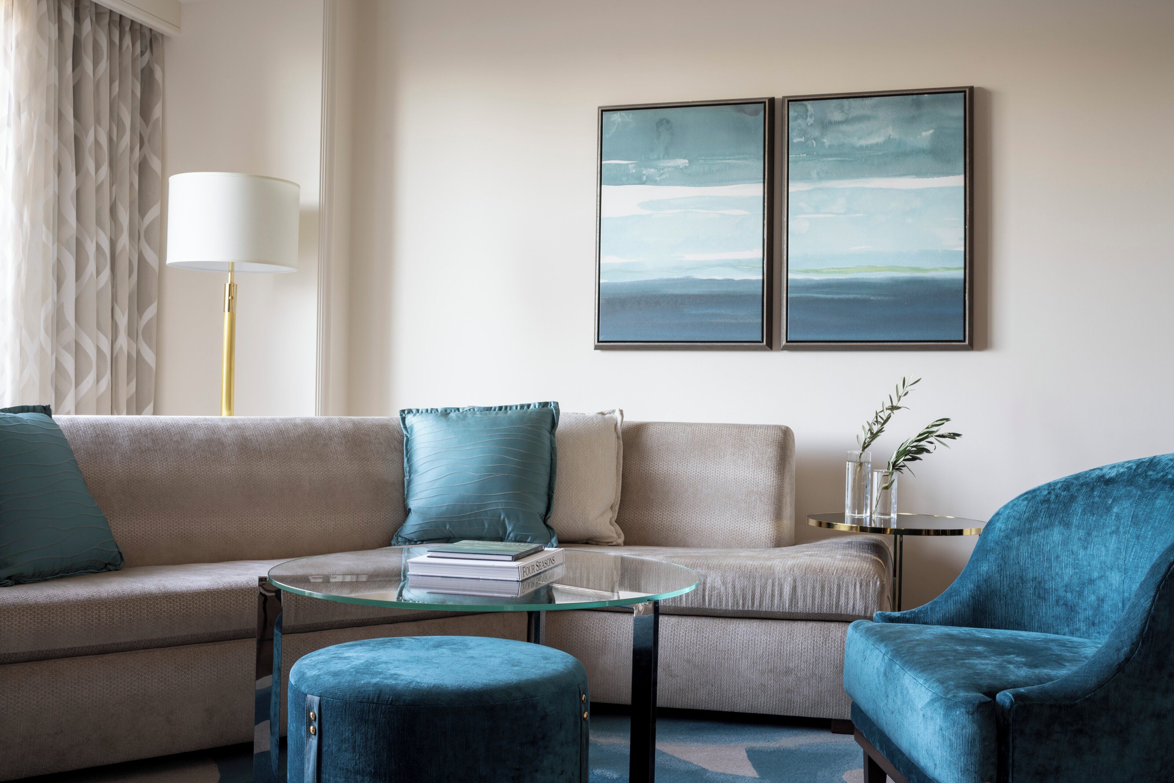 A modern living room with a beige sofa, a glass coffee table, a blue armchair, and two framed abstract paintings on the wall.