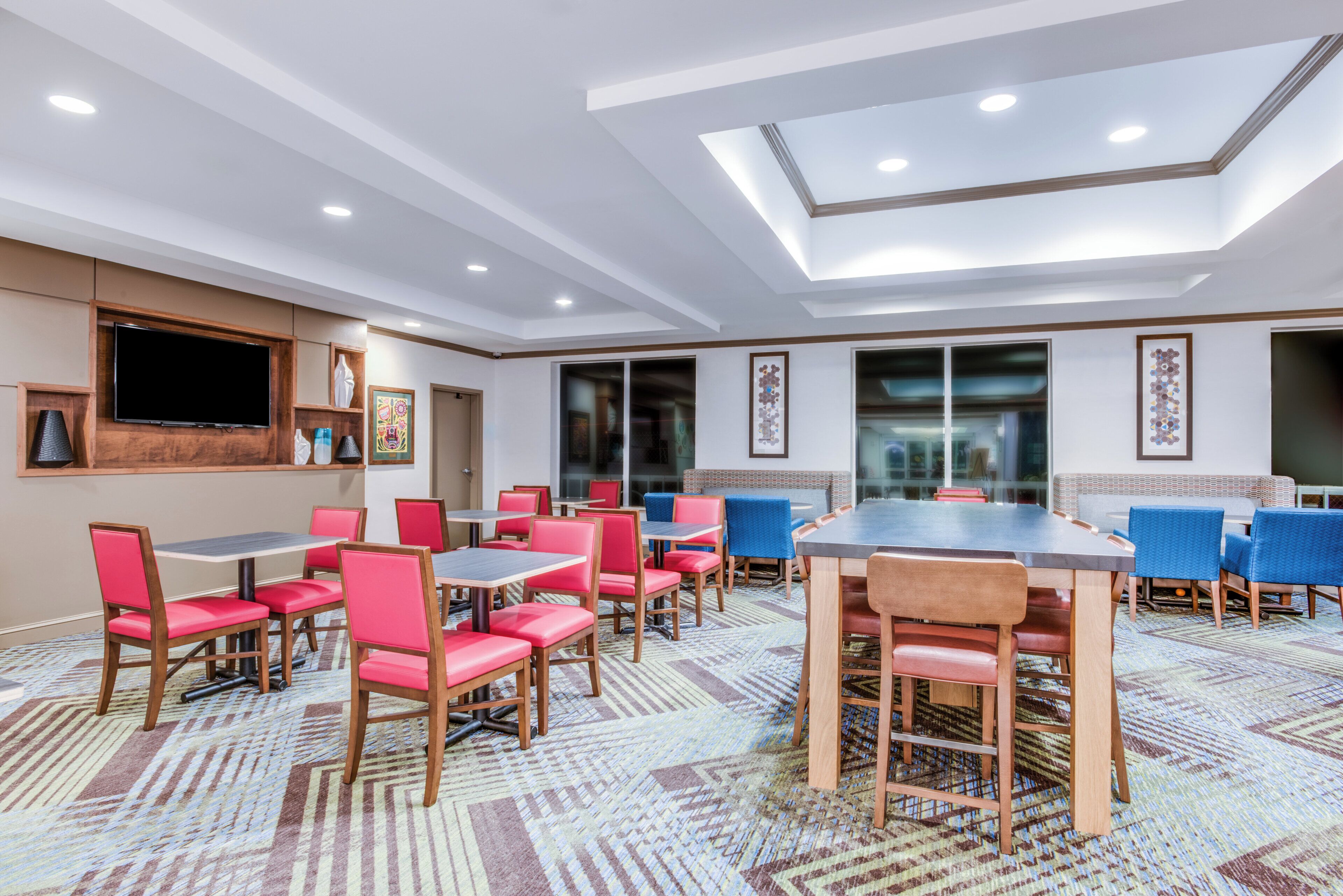 Un comedor de hotel con varias mesas y sillas, un televisor de pantalla plana montado en la pared y una alfombra estampada.