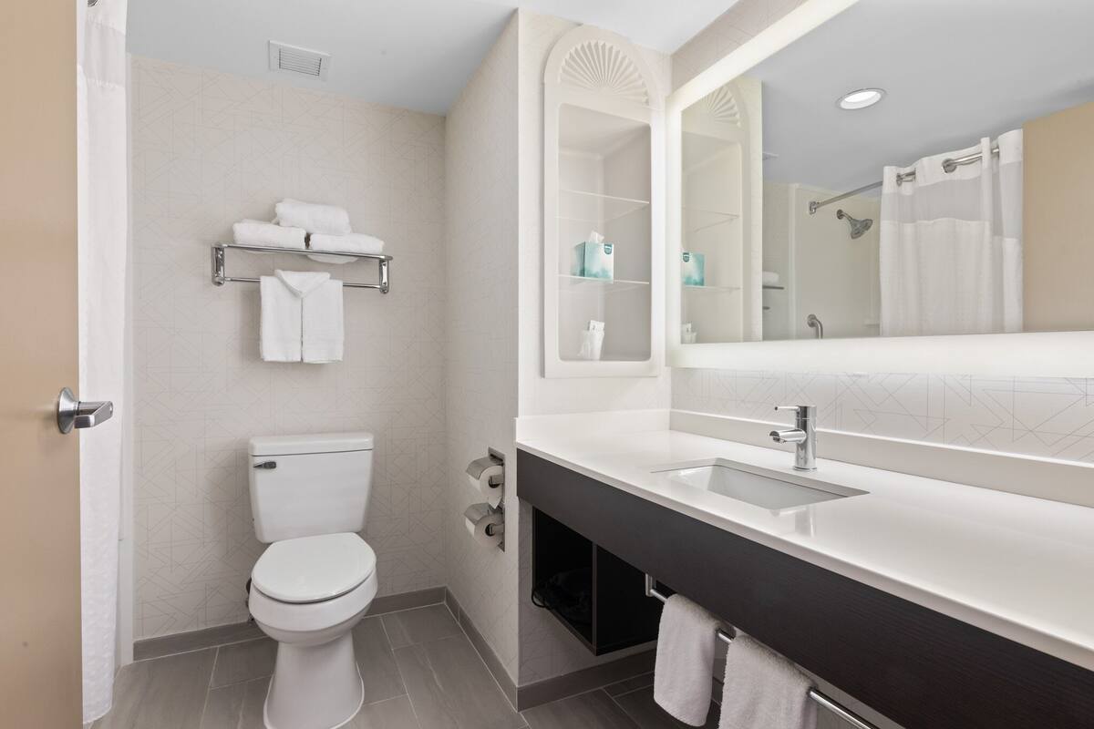 A modern bathroom with a toilet, sink, and mirror. There are towels on the wall and a shelf with toiletries.