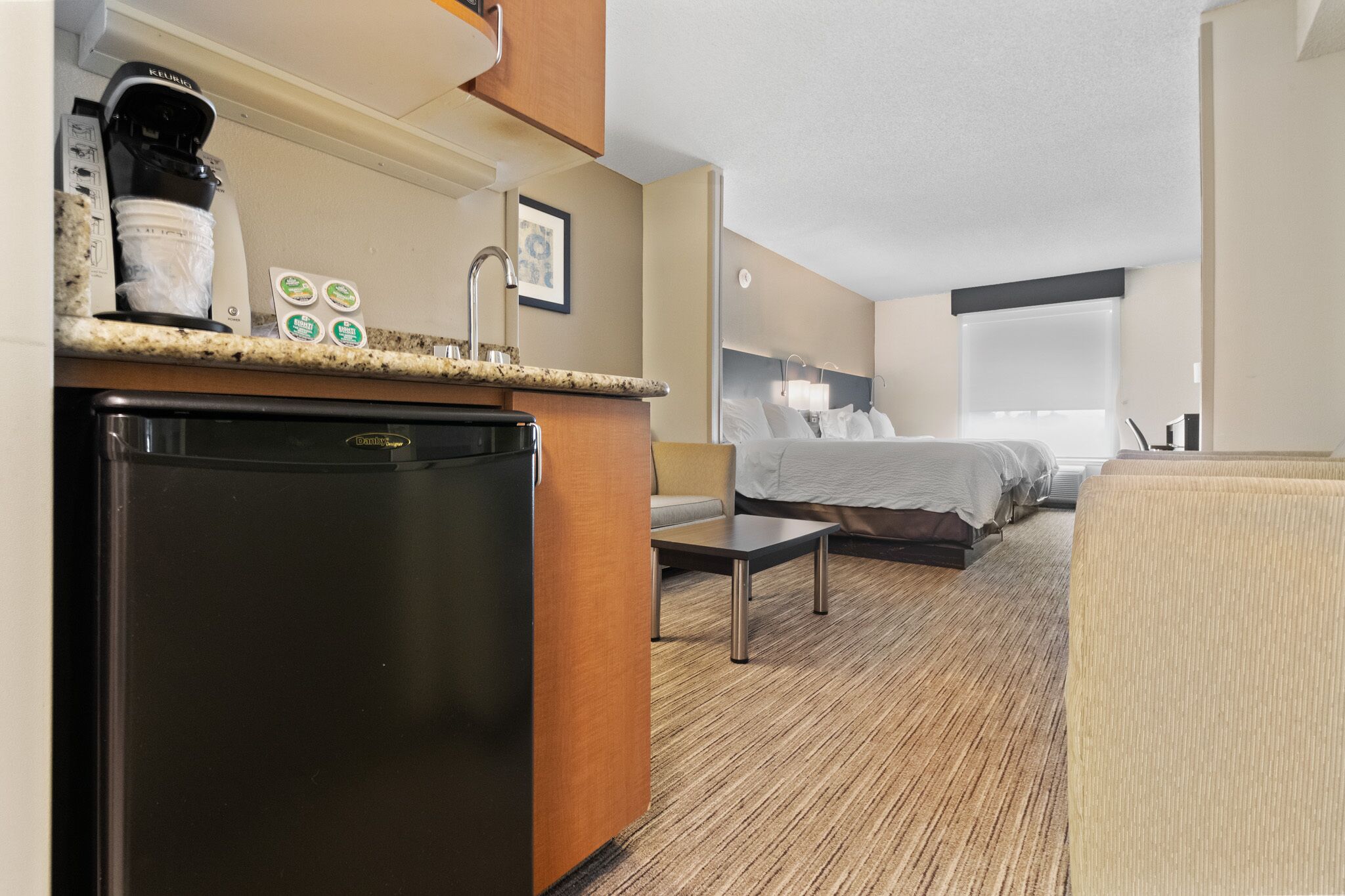 Quarto de hotel com cozinha compacta, cama, mesa de cabeceira e janela.