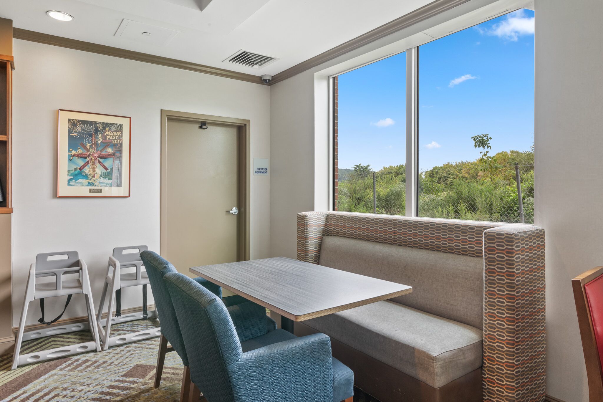 Habitación con un banco, una mesa y dos sillas. Se ve una puerta y una ventana con vistas a árboles.