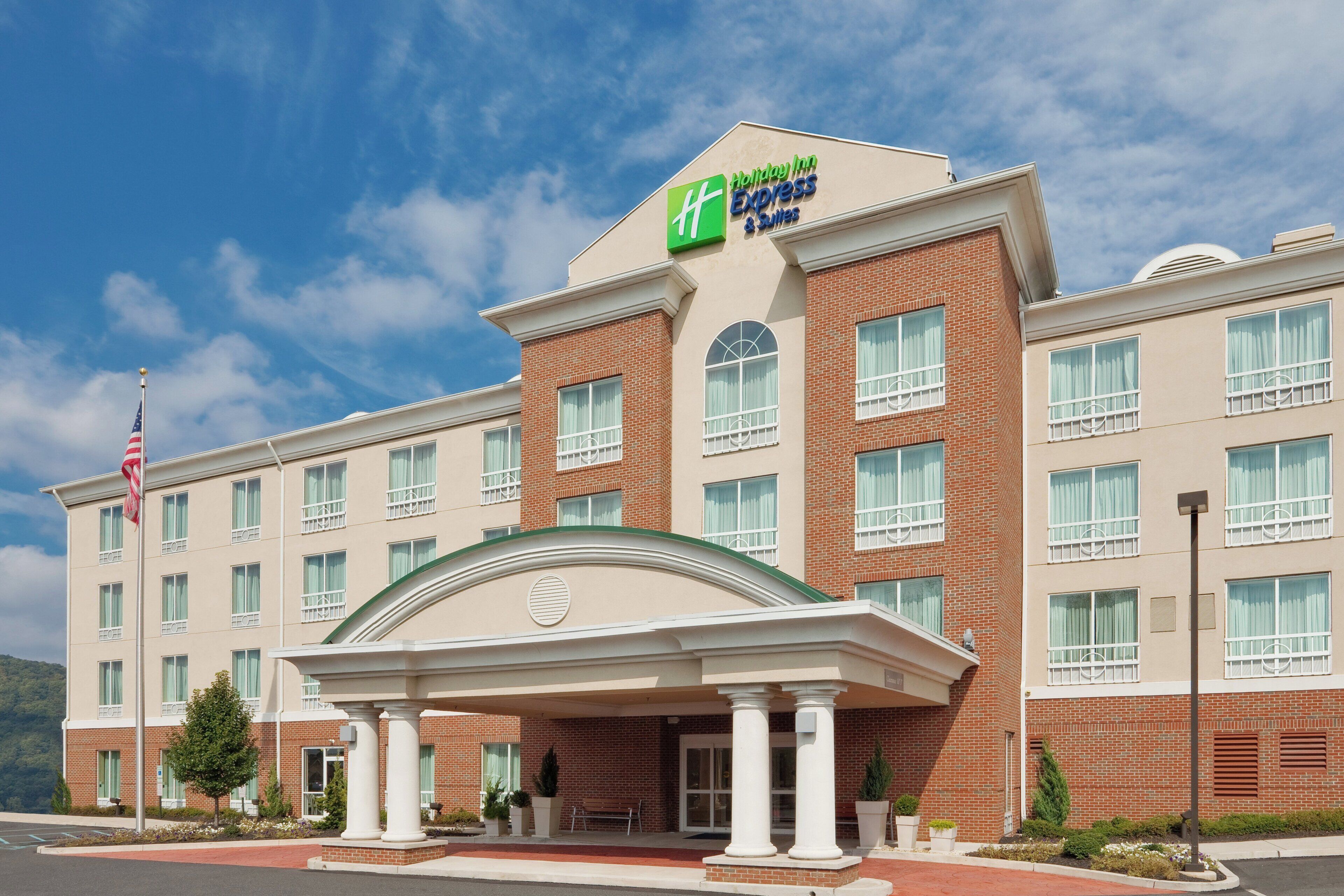 Um hotel Holiday Inn Express com fachada de tijolos, entrada central com varanda coberta e um mastro com a bandeira americana.