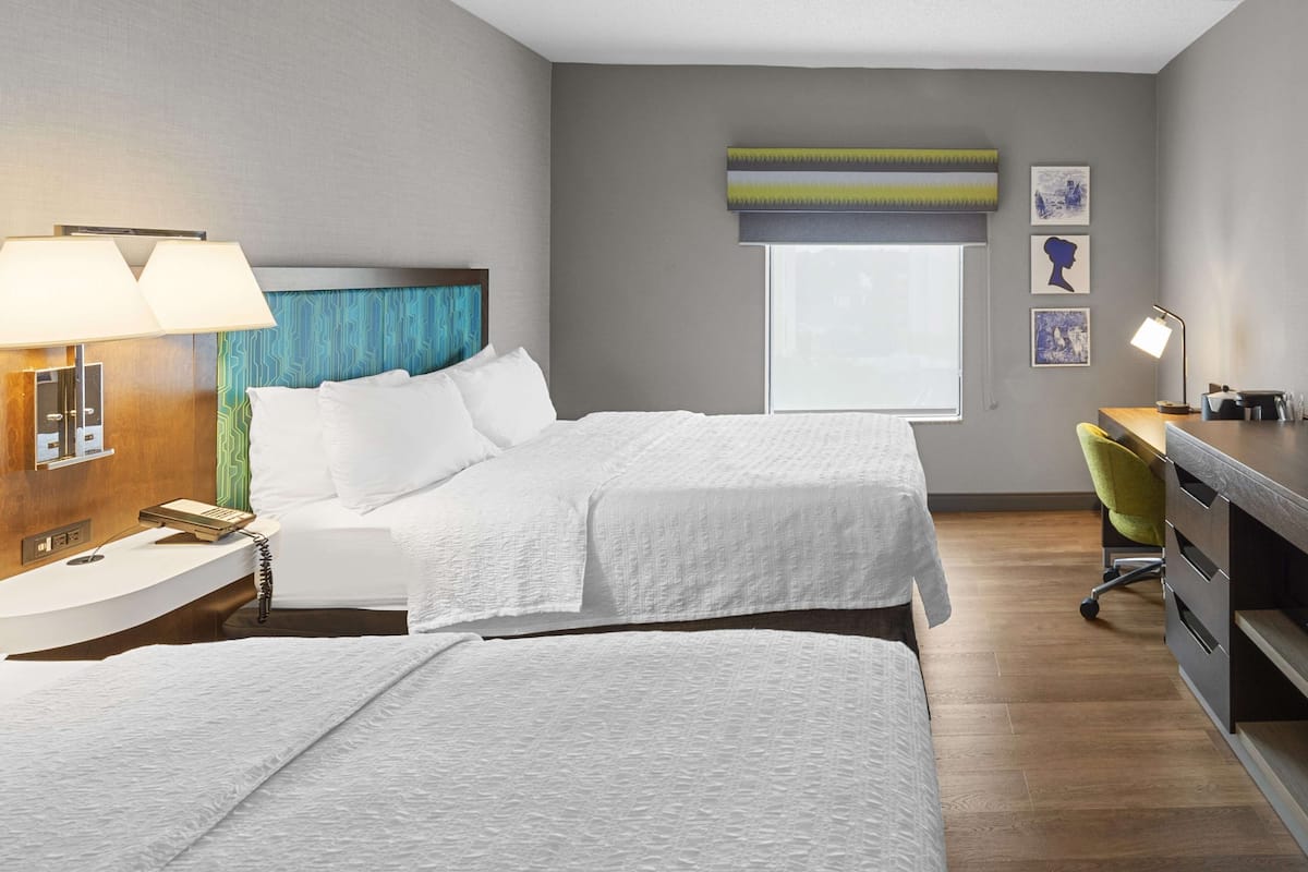 A hotel room with two beds, a desk, a chair, a lamp, and artwork on the wall.
