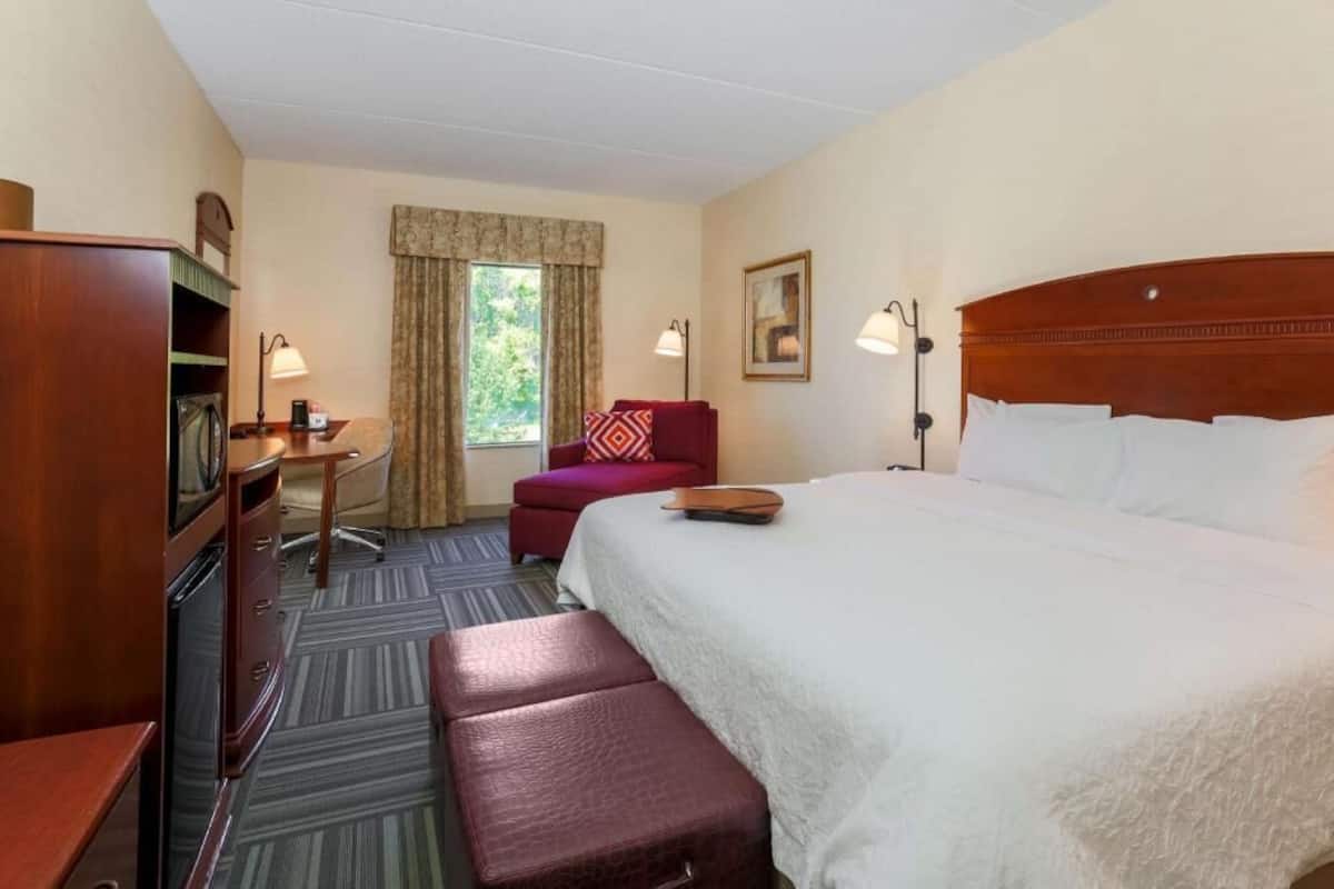 A hotel room with a bed, a desk, a chair, a bench, a lamp, a picture frame, and a window with curtains.