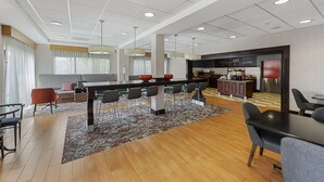A modern hotel lobby with a long communal table, seating area, and a kitchenette.
