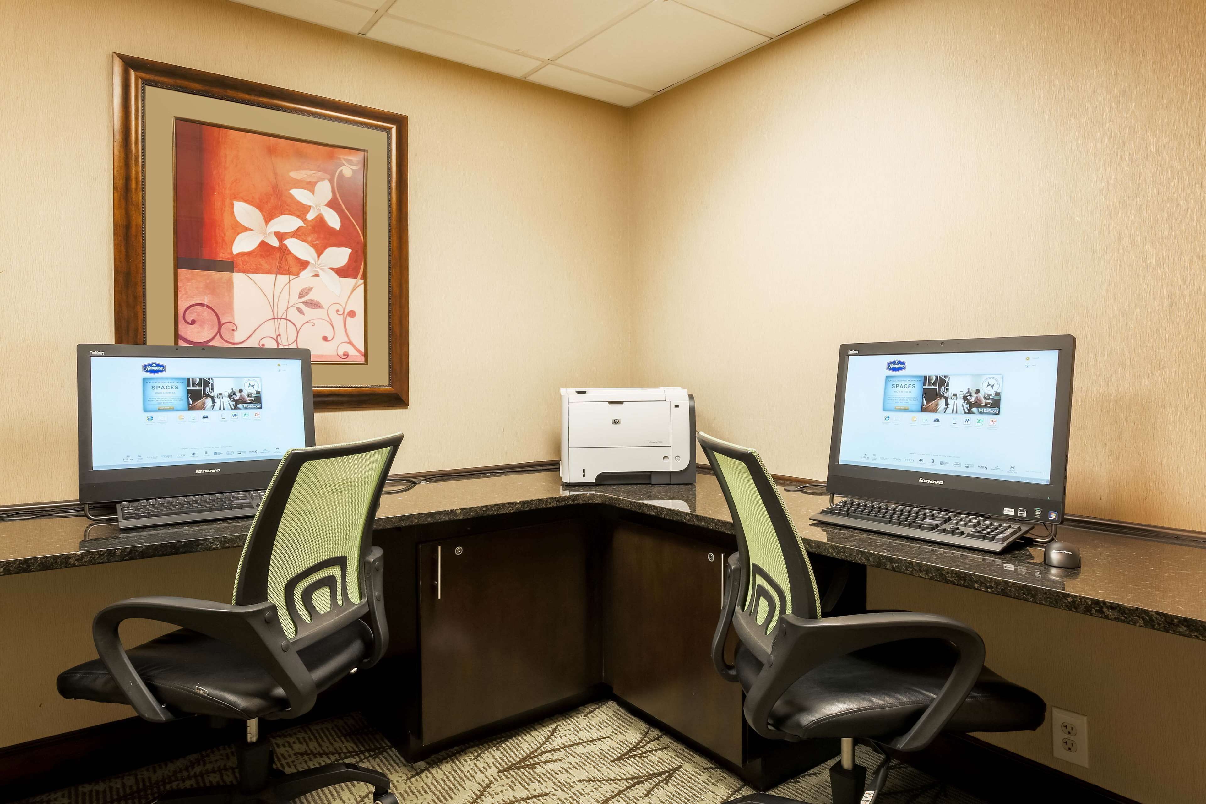 A hotel workspace with two desks, each with a computer and chair, a printer, and a framed artwork on the wall.