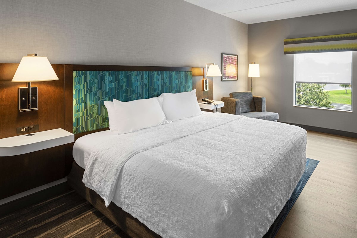 A hotel room with a large bed, a nightstand, a chair, a desk, and a window with a view.