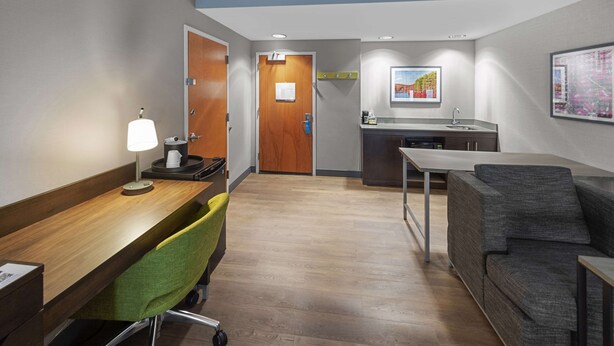 A hotel room with a wooden desk, a green chair, a gray sofa, a kitchenette, and a door with a sign.