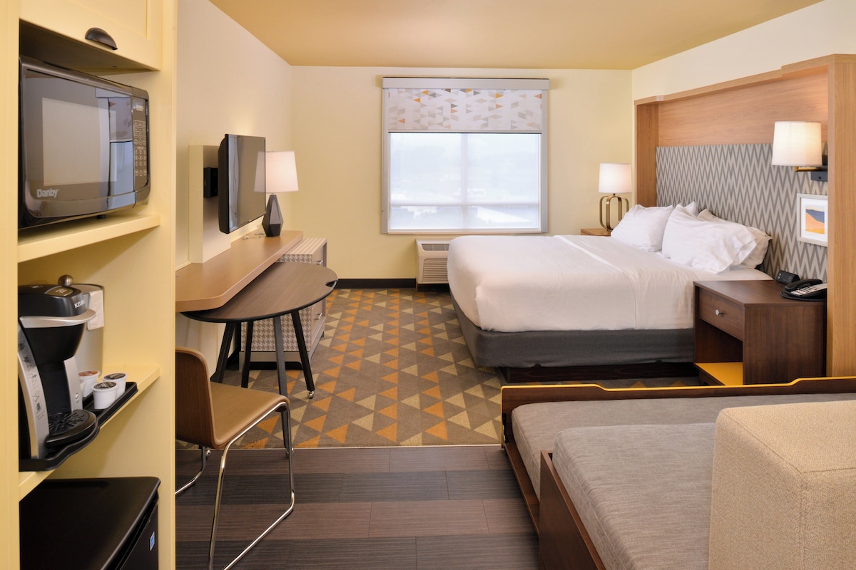 Quarto de hotel com cama, escrivaninha com cadeira, televisão e janela com persiana estampada.