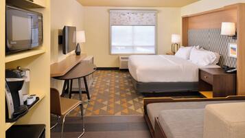 A hotel room with a bed, a desk with a chair, a television, and a window with a patterned blind.