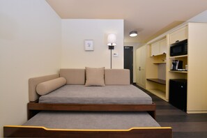 Quarto compacto com cama, luminária de cabeceira, pequena escrivaninha e área de cozinha compacta.