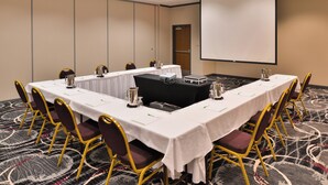 Un salón de conferencias con una larga mesa, sillas, una pantalla de proyector y piso alfombrado.