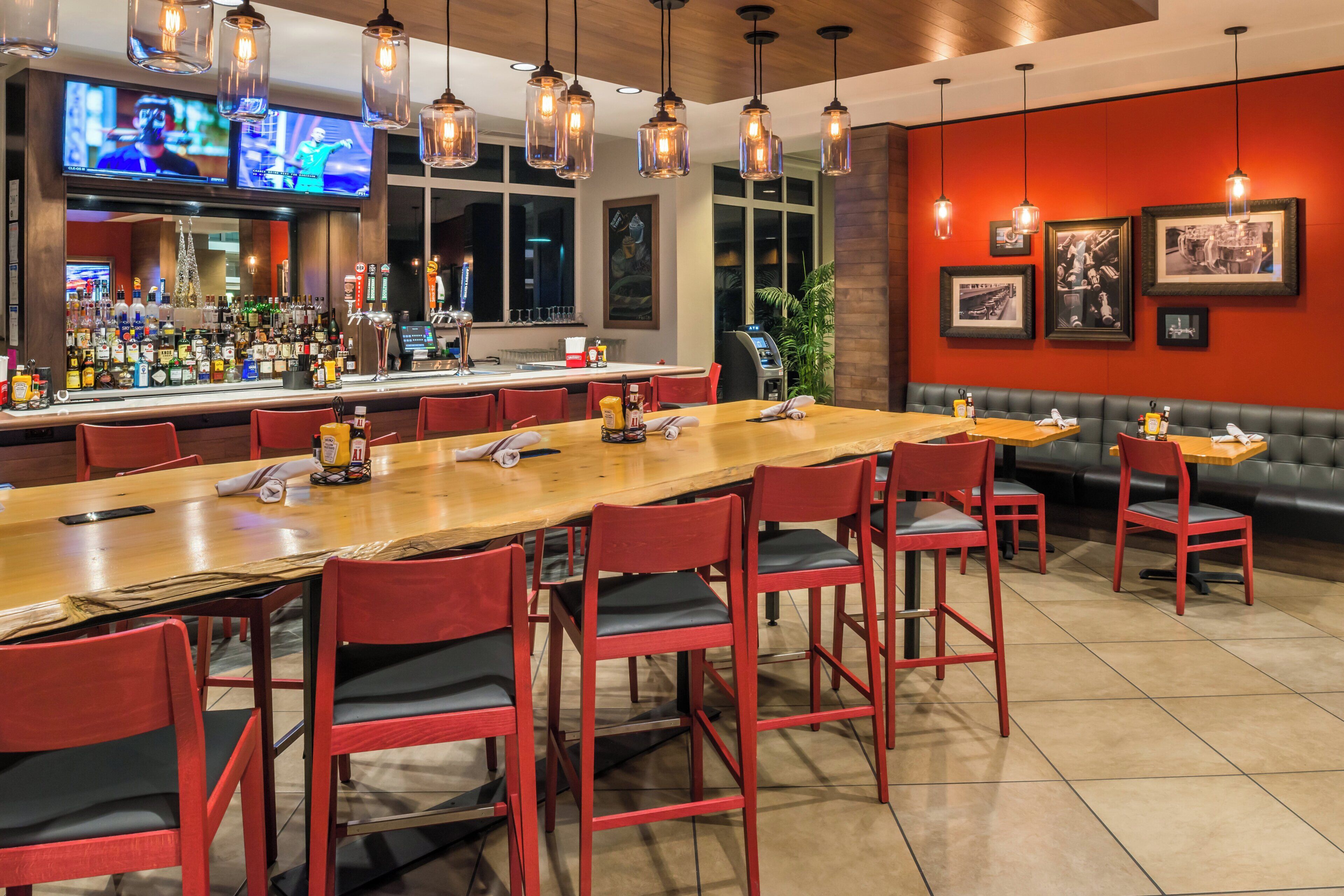 A bar area with a long wooden counter, red chairs, and hanging light fixtures.