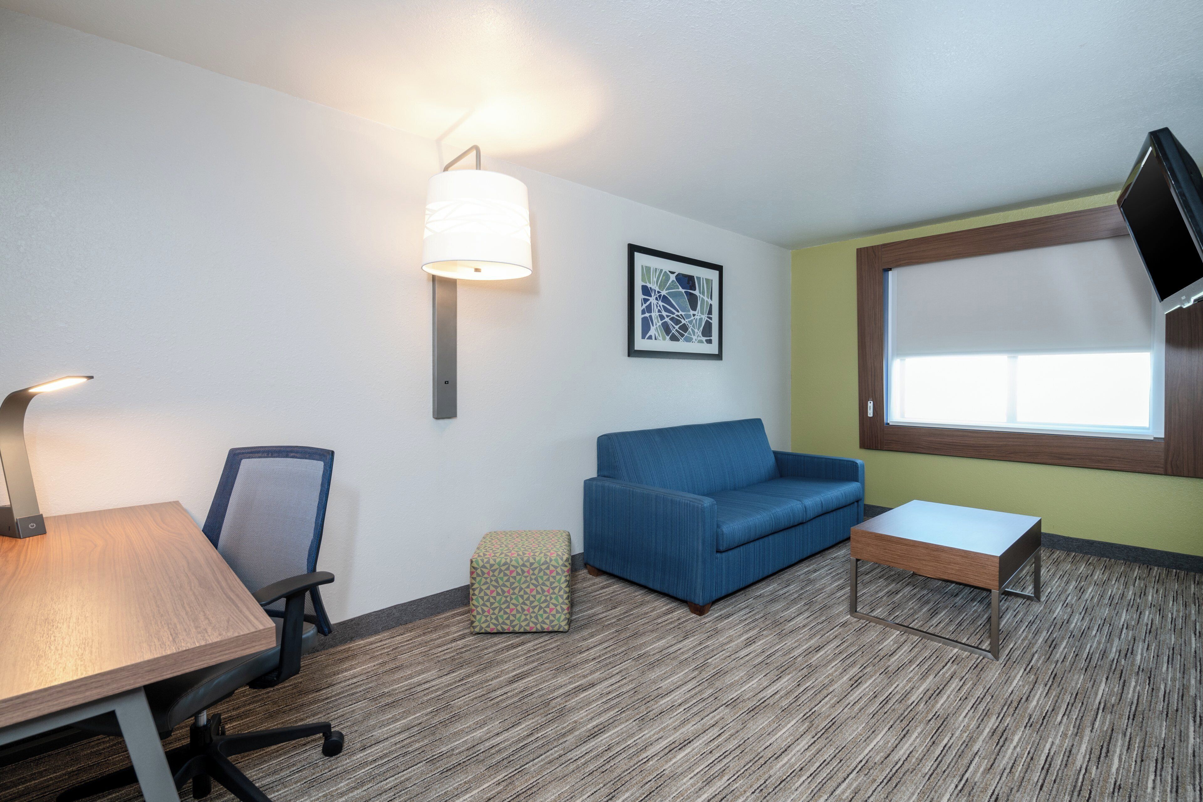 A hotel room with a desk, chair, sofa, ottoman, and a wall-mounted TV.