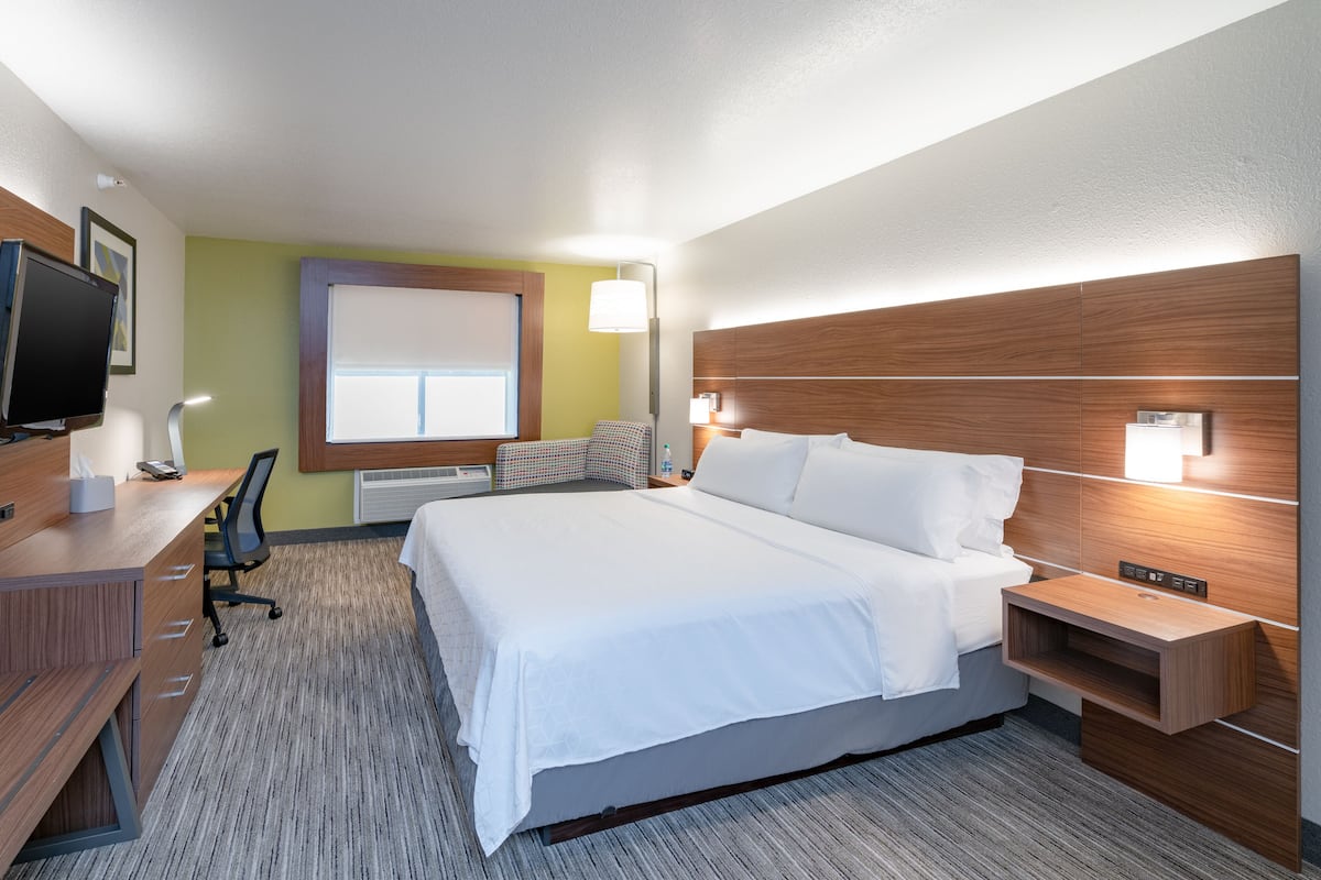A hotel room with a large bed, a desk with a computer, a chair, a TV, and a window with blinds.