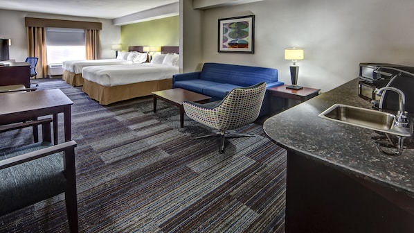 Quarto de hotel com duas camas, um sofá azul, uma poltrona pequena, uma mesa de trabalho e uma kitchenette.