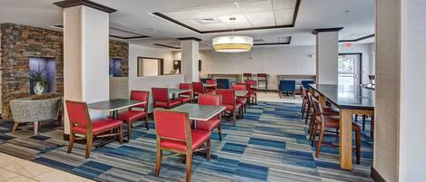 A hotel dining area with multiple tables and chairs, a patterned carpet, and a stone accent wall.