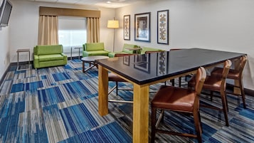 Una habitación de hotel con una amplia mesa de conferencias, sofás verdes, una mesita auxiliar y una lámpara.