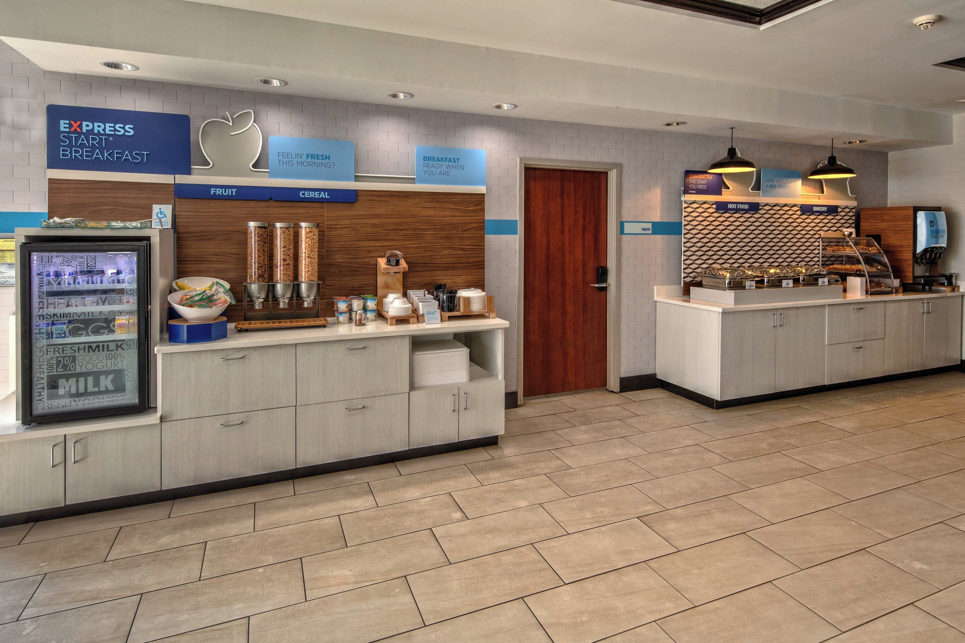 A hotel breakfast area with a self-serve station, a refrigerator with fresh milk and yogurt, and a bakery section.