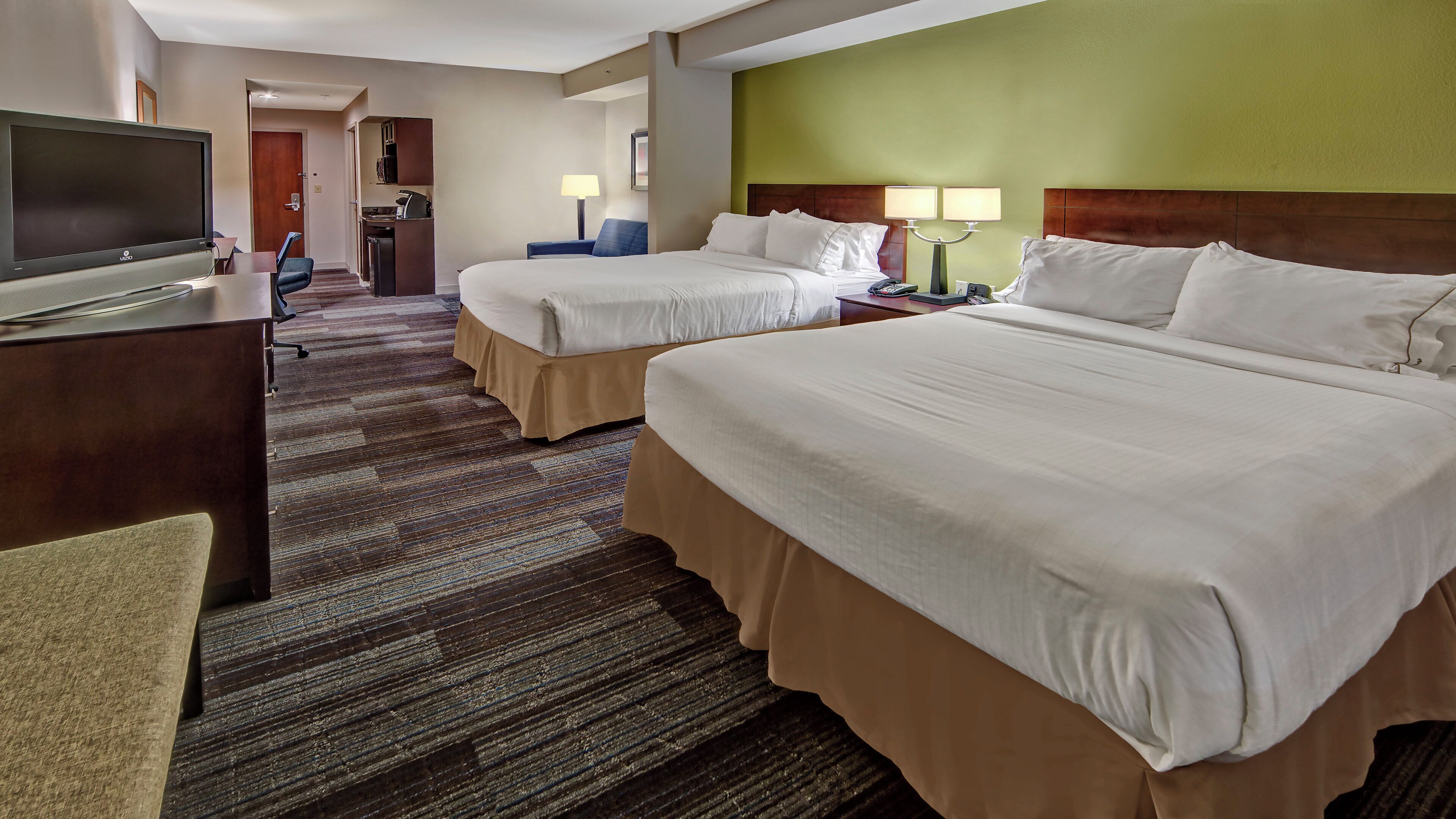 A hotel room with two beds, a desk with a TV, and a kitchenette.