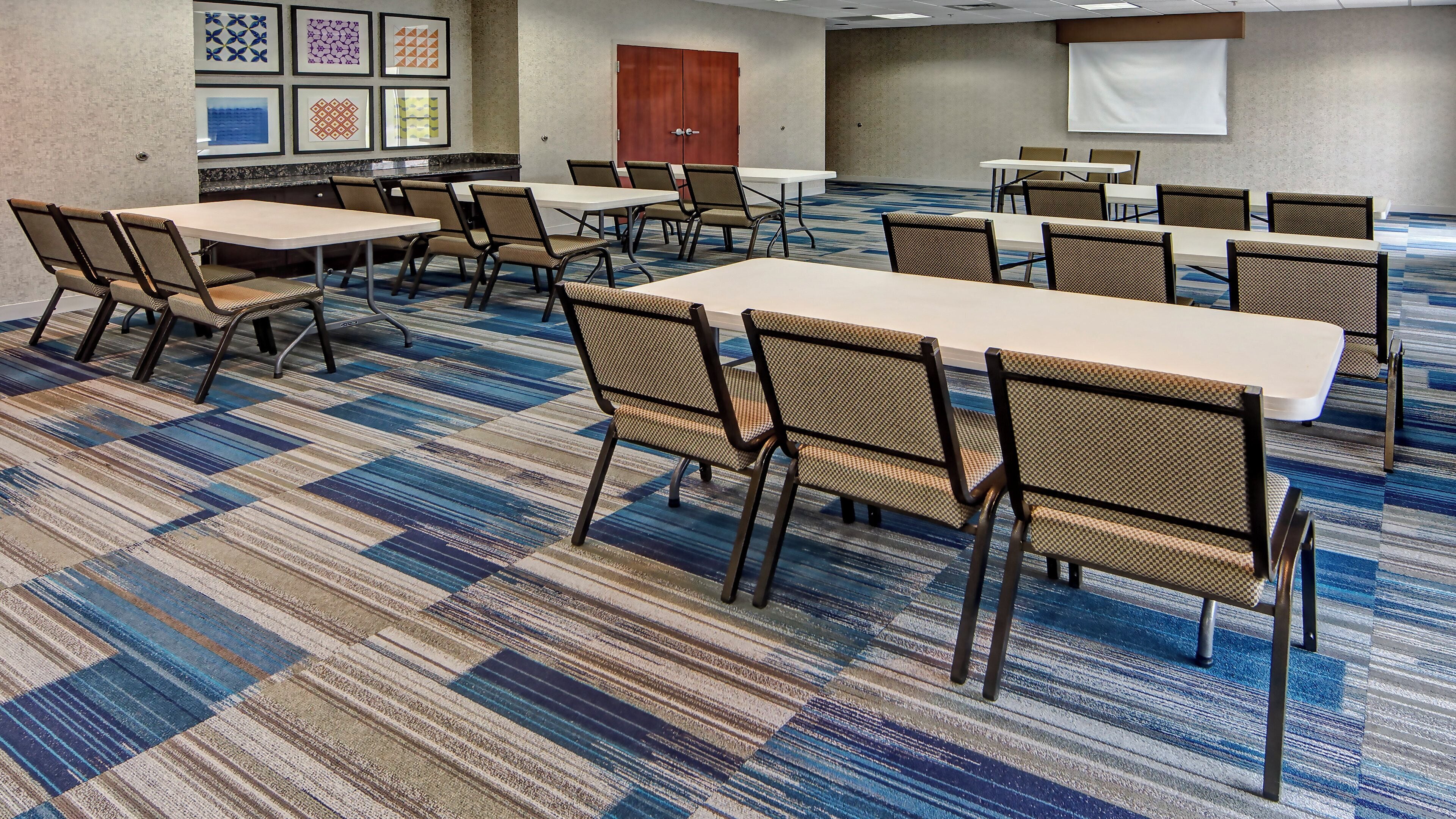 A conference room with tables and chairs, a projector screen, and framed artwork on the wall.