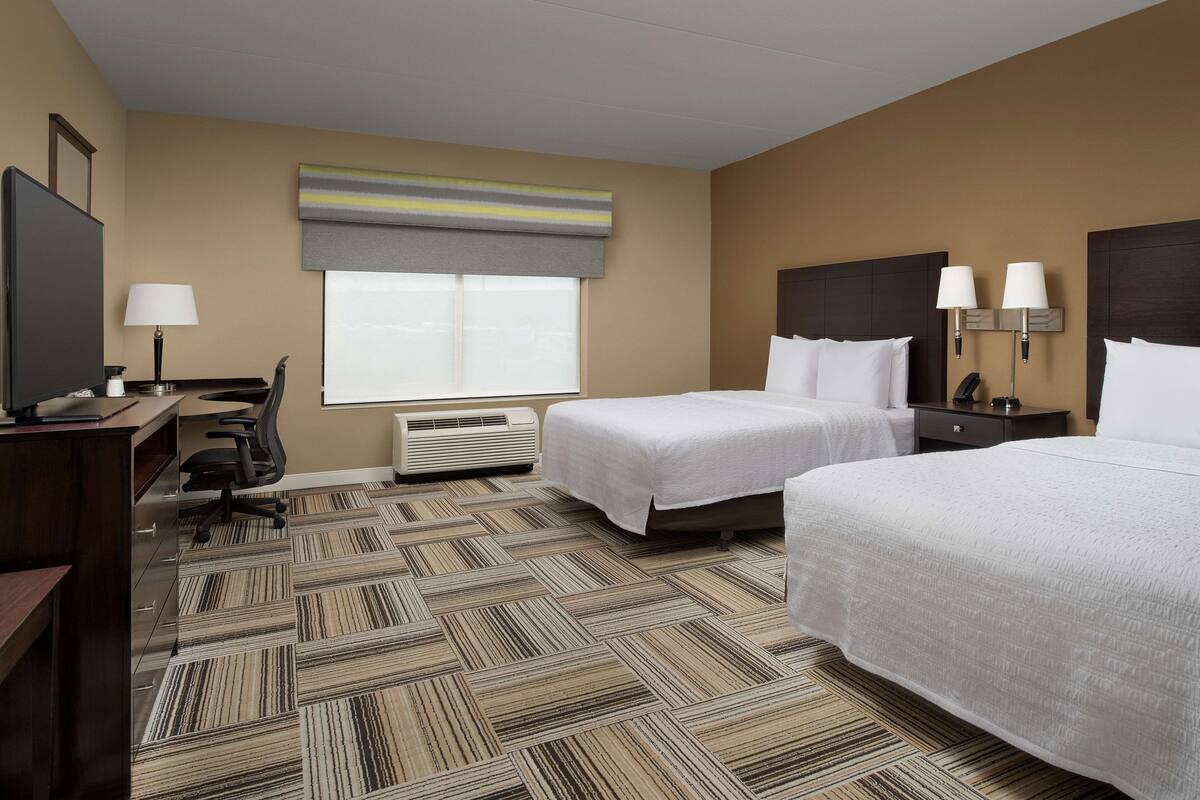 A hotel room with two beds, a desk with a chair, a TV, and a window with a striped blind.