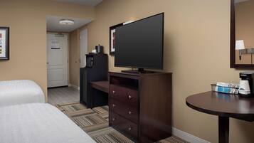 A hotel room with two beds, a desk with a flat-screen TV, a coffee maker, and a small table with a basket of items.