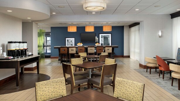 A hotel dining area with multiple tables and chairs, a coffee station, and a television screen. - Hampton Inn Knoxville East (Knoxville)