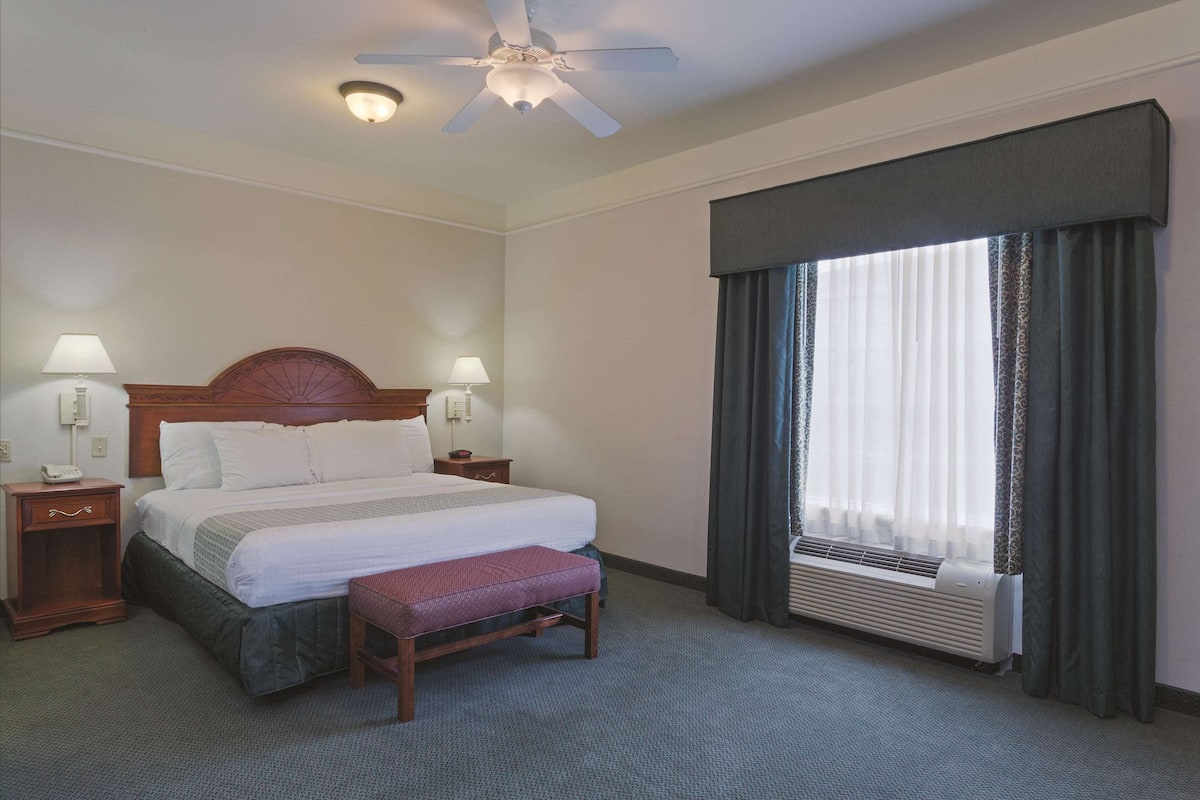 Une chambre d’hôtel comprenant un lit, deux tables de chevet avec lampes, un ventilateur de plafond, une fenêtre avec rideaux et un système de climatisation intégré.