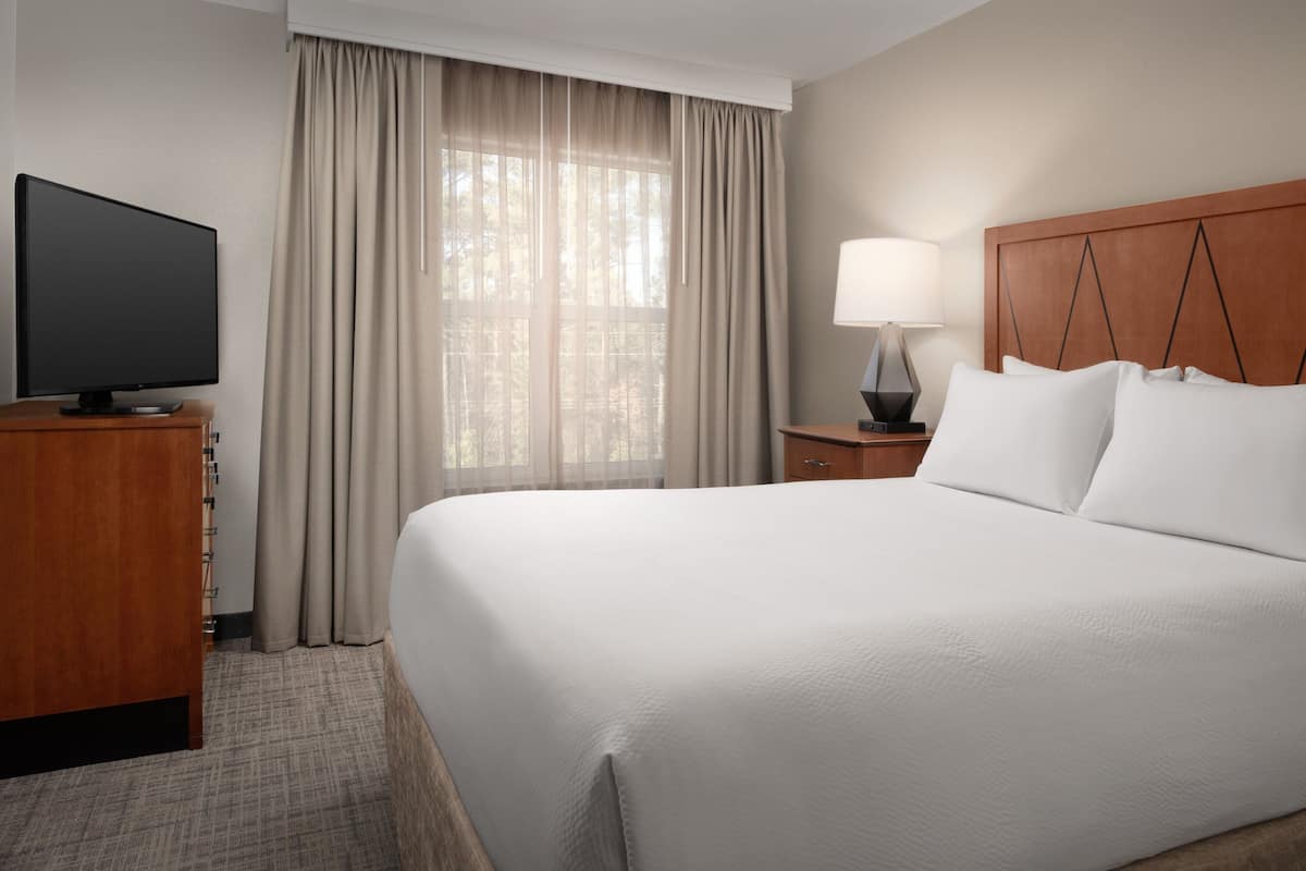 Habitación de hotel con una cama grande, un televisor sobre un soporte de madera, una mesita de noche con lámpara y una ventana con cortinas.
