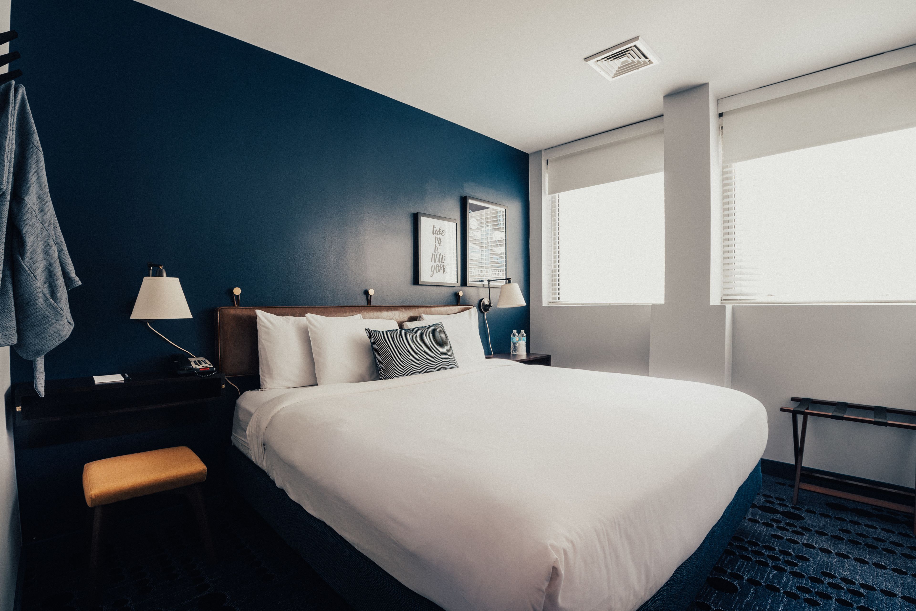 A hotel room with a large bed, a bedside table with a lamp, a small stool, a wall-mounted towel rack, and a window with blinds.